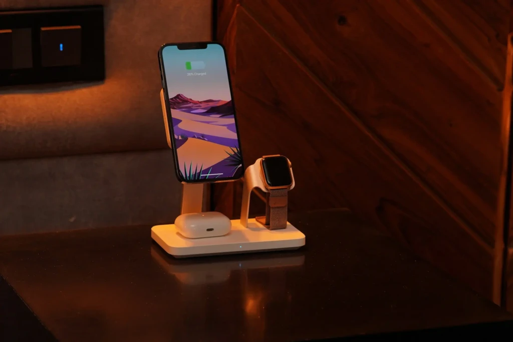 A white multi-device wireless charger on a dark wooden nightstand holding an iPhone (displaying “26% Charged” and a desert wallpaper), an Apple Watch with a woven band, and an AirPods case, illuminated by warm ambient lighting with a smart light switch visible in the background.