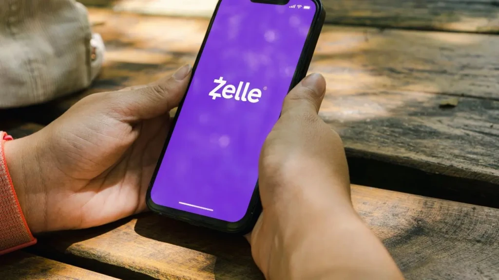 A person holds a smartphone displaying the Zelle app on a purple background, with a wooden table in the background.