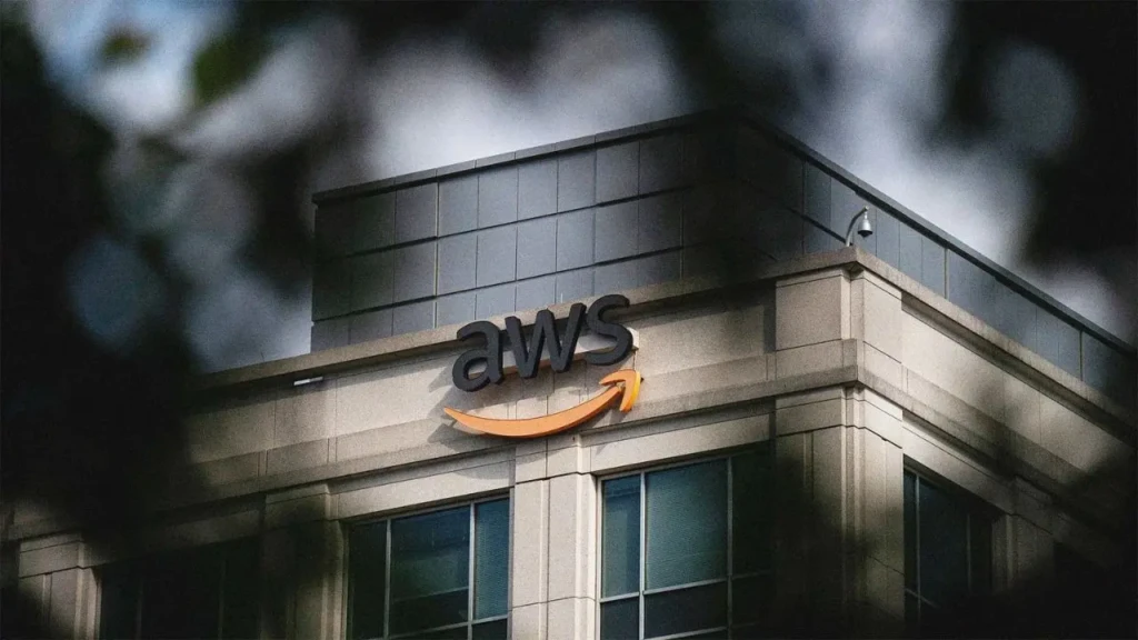 The AWS logo mounted on the exterior corner of a modern office building, partially framed by out-of-focus tree leaves in the foreground, conveying the physical presence and corporate identity of Amazon Web Services in an urban setting.