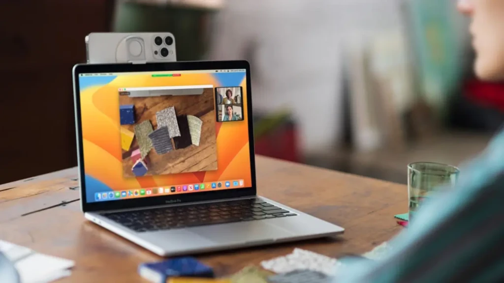 A MacBook Pro on a wooden desk with an iPhone mounted on top via a stand, displaying a FaceTime video call with multiple participants; the laptop screen shows a design app with fabric swatches, illustrating seamless Continuity and cross-device collaboration in macOS Ventura.
