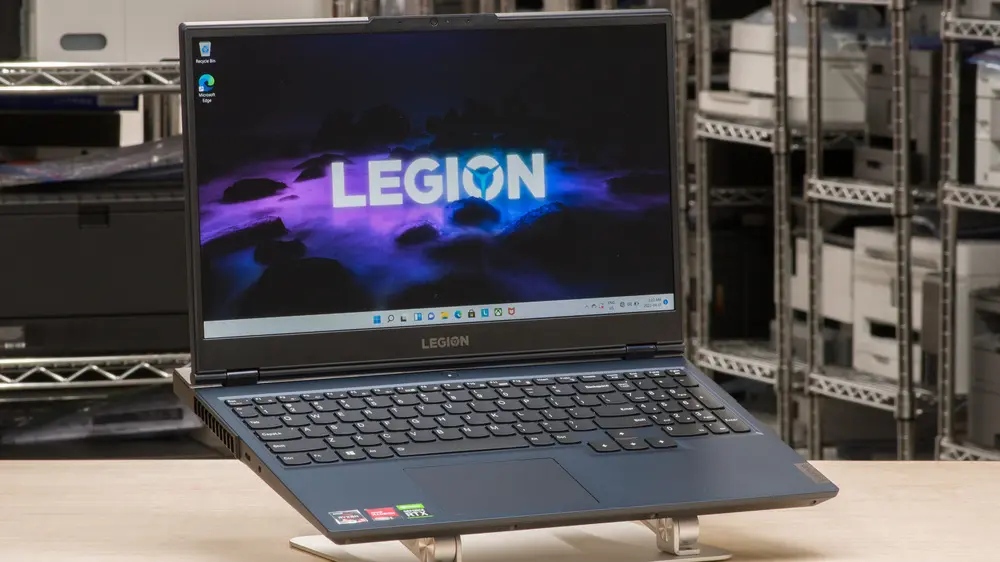A dark-blue Lenovo Legion laptop on a stand in a warehouse-like environment, showing the “LEGION” logo wallpaper on screen, Windows desktop icons, and visible spec stickers (RTX, Intel), emphasizing its professional-grade build and readiness for productivity or gaming in a tech-focused setting.