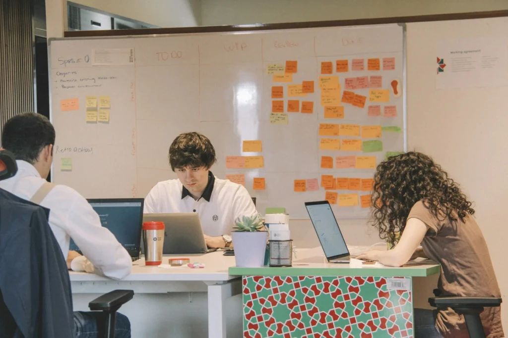 A group working collaboratively in an office setting, with laptops open and a whiteboard filled with colorful sticky notes.