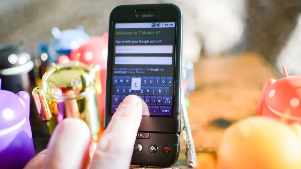 A close-up of a hand typing on the touchscreen keyboard of a black T-Mobile G1 during Google account sign-in, surrounded by colorful Android robot figurines, symbolizing the device’s launch era and its foundational role in the Android ecosystem.