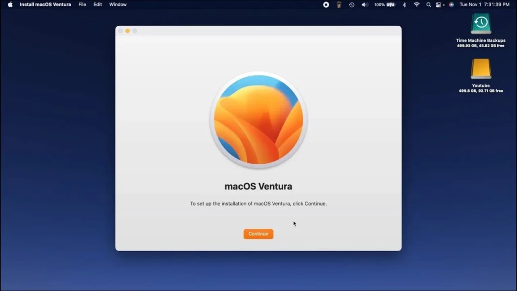 The macOS Ventura installer window on a Mac desktop, displaying the orange-and-blue floral logo, the text “macOS Ventura,” and an orange “Continue” button, with system status icons (Time Machine, YouTube) visible in the top-right corner, indicating the initial setup phase of the operating system installation.
