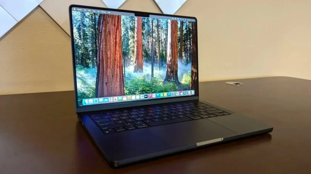 A space-black MacBook Pro angled on a dark wooden desk, showing a redwood forest wallpaper, macOS menu bar, and dock, emphasizing its immersive screen, sleek profile, and professional workspace integration.