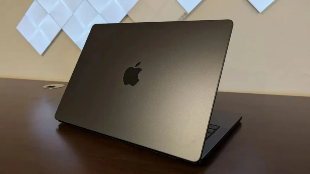 A matte space-black MacBook Pro closed and propped open slightly on a dark wood desk, with the Apple logo centered on the lid and geometric wall art in the background, showcasing its minimalist design and premium finish in a curated studio environment.