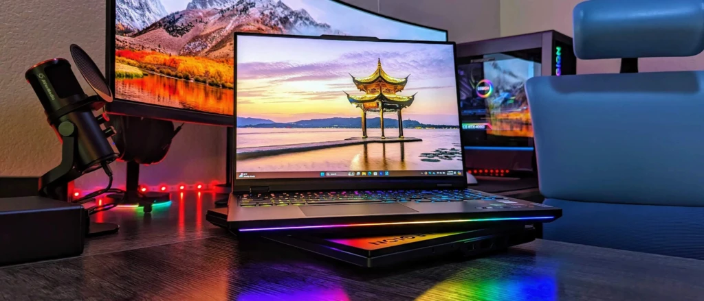 A high-end gaming desk setup featuring a Lenovo Legion laptop with rainbow RGB keyboard and chassis lighting, positioned between a large curved monitor (showing a scenic pagoda wallpaper) and a transparent PC tower with RGB fans, alongside an Audio-Technica microphone, showcasing a complete, premium creator/gamer ecosystem.
