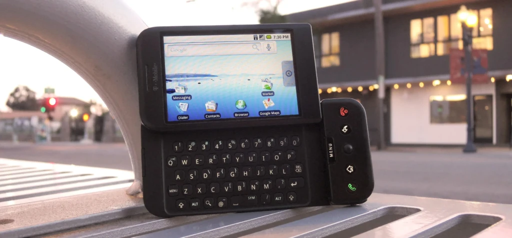 A black T-Mobile HTC Dream placed on an outdoor metal bench at dusk, displaying its home screen with Google search and app icons, while city lights and traffic signals blur in the background, illustrating its real-world usability and historic presence in early mobile computing.