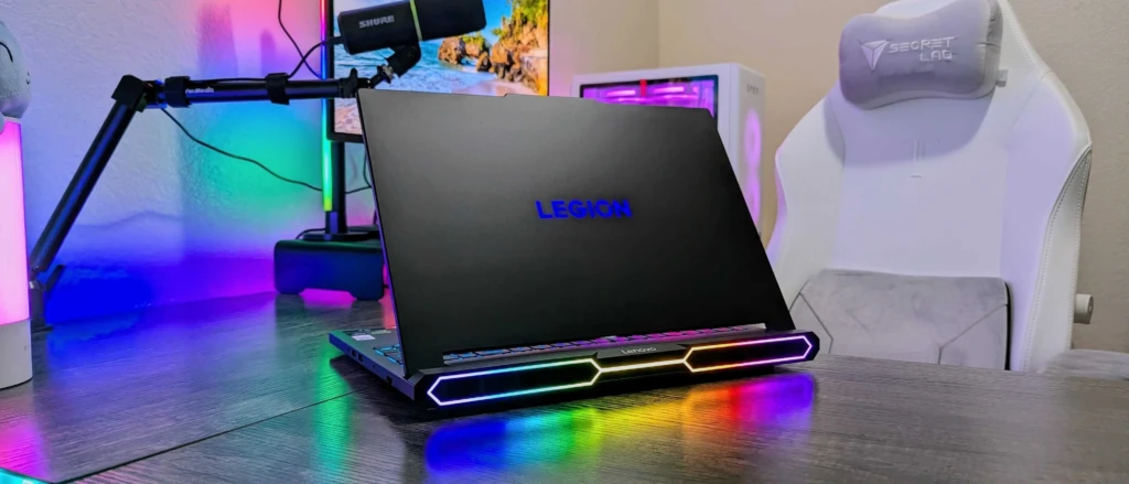 A sleek Lenovo Legion gaming laptop with RGB lighting sits on a desk beside a white gaming chair, with colorful ambient lighting in the background.