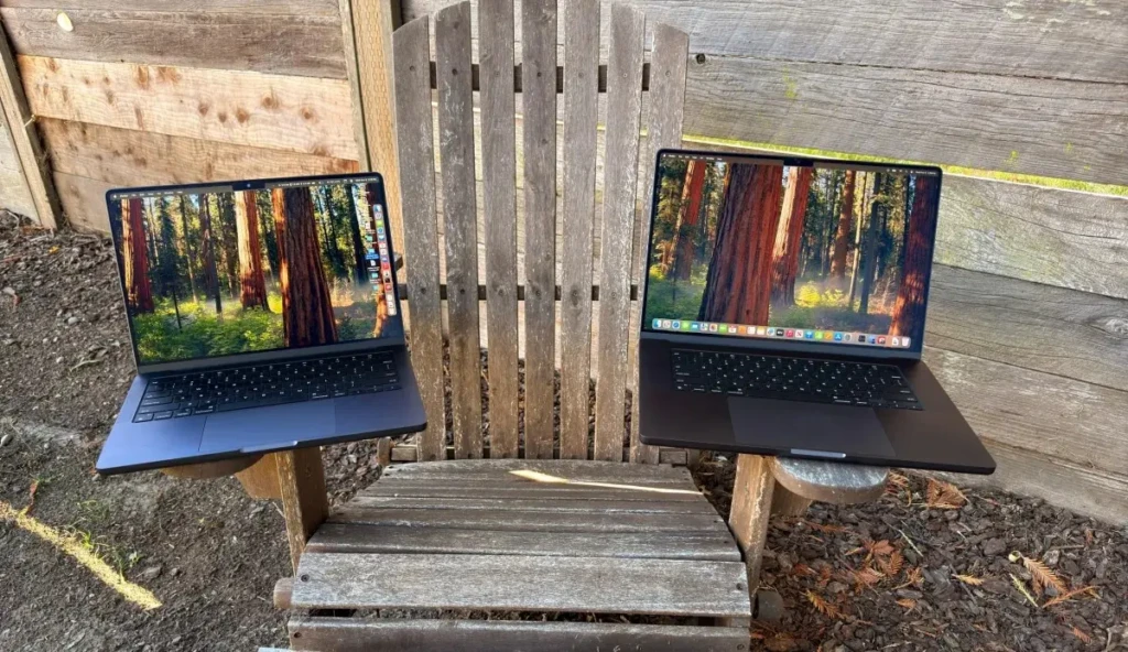 Two identical space-black MacBook Pros placed side by side on the armrests of a weathered wooden Adirondack chair against a rustic fence backdrop, both displaying the same redwood forest wallpaper, illustrating real-world portability, symmetry, and outdoor usability.