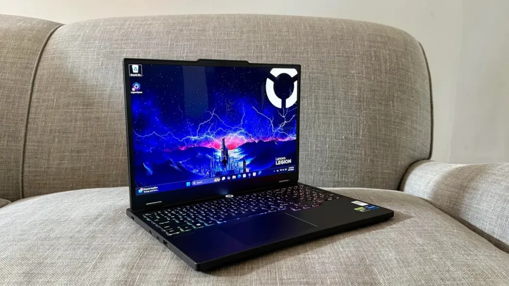 A black Lenovo Legion laptop open on a gray fabric sofa, displaying a dynamic cosmic-and-cityscape wallpaper with the LEGION logo and RGB-backlit keyboard, highlighting its portability and adaptability for casual or lounge-based gaming and content creation.