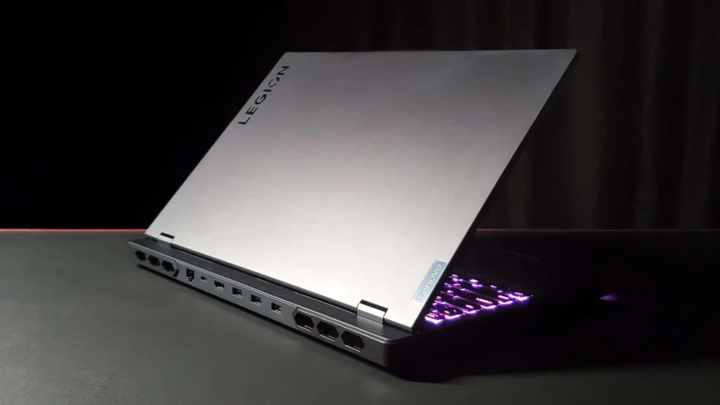 A silver Lenovo Legion laptop angled to reveal its left-side I/O panel with multiple ports (USB-A, HDMI, Ethernet, etc.), while the lid displays the “LEGION” branding and the keyboard glows with purple RGB backlighting, emphasizing connectivity, build quality, and aesthetic customization against a dark backdrop.