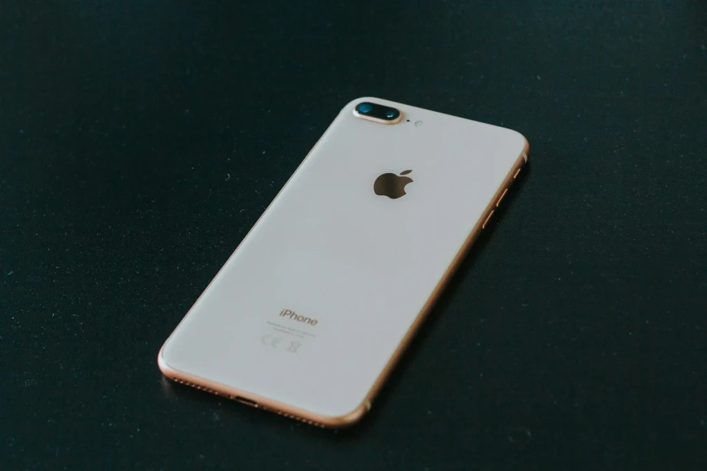 A rose gold iPhone 8 lying diagonally on a dark textured surface, showing its glossy white back panel, dual-camera module, Apple logo, and “iPhone” branding, highlighting its classic design and metallic finish in a minimalist studio shot.