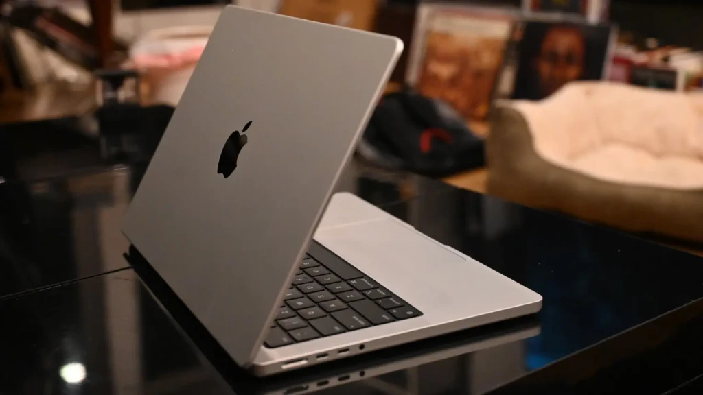 A silver MacBook Pro partially open on a glossy black table, with the Apple logo visible on the lid and ports (USB-C, headphone jack) along the left edge, set against a softly blurred indoor background featuring books and decor, conveying a modern home-office aesthetic.