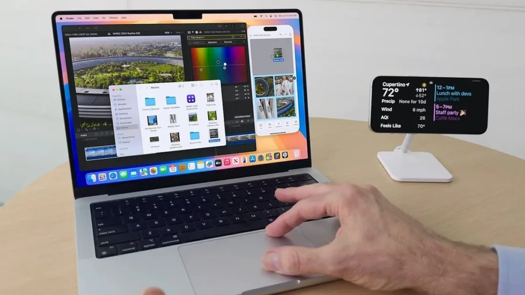 A person using a MacBook with multiple windows open, including Final Cut Pro, Finder, and a color picker, while a nearby iPhone on a stand displays a smart home widget with weather, calendar, and AQI data; the user’s hand rests on the trackpad, highlighting seamless multitasking and ecosystem integration in macOS Sequoia.