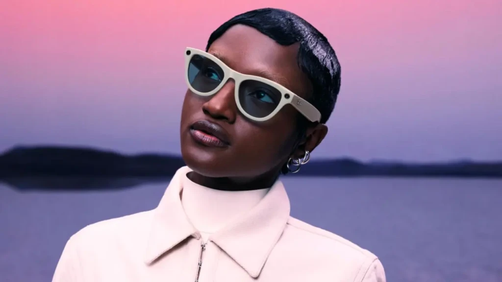 A person with short dark hair wearing cream-colored Ray-Ban Meta smart glasses with dark lenses, set against a dreamy pink-to-purple sunset over water, showcasing the product in a stylish, lifestyle-oriented context with visible camera modules on the temples.