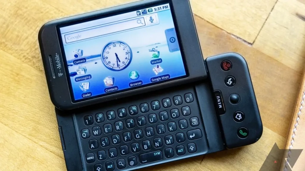 A black T-Mobile HTC Dream resting on a wooden surface, with its screen showing the Google search bar, clock widget, and app icons including Messaging, Browser, and Google Maps, emphasizing its early Android interface and physical navigation controls on the side panel.