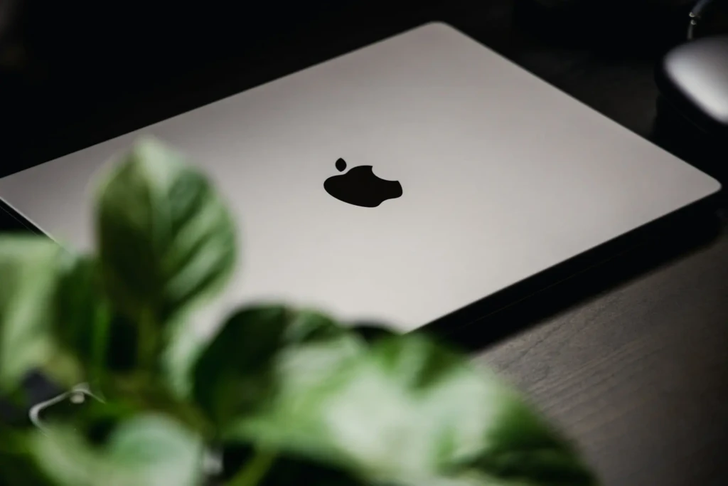 A closed silver MacBook Pro resting on a dark wooden surface, with the black Apple logo centered on the lid and blurred green foliage in the foreground, evoking a clean, modern workspace aesthetic and understated sophistication.