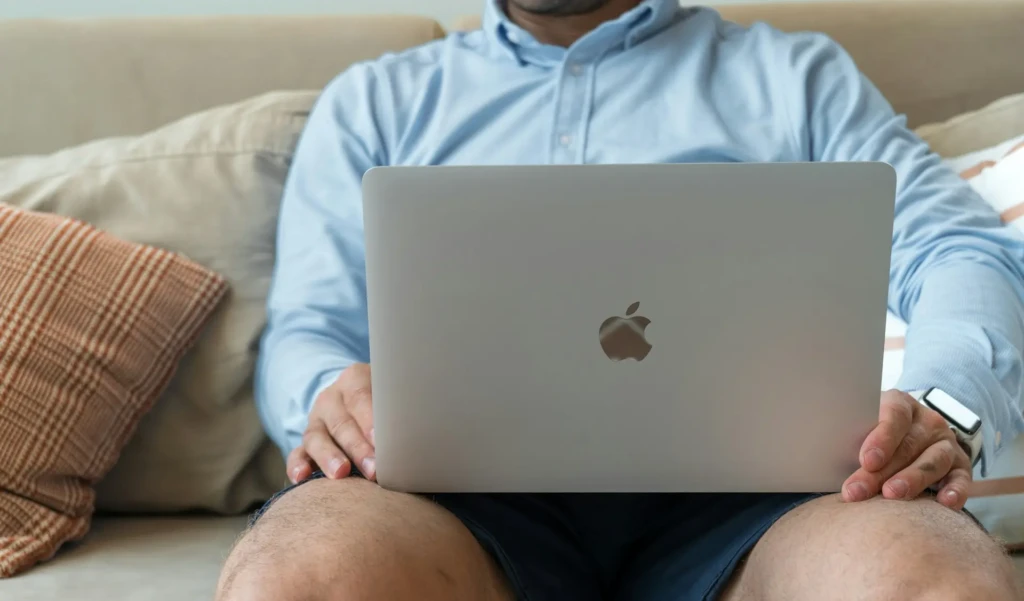 A person in a light blue shirt sitting on a couch with a silver MacBook on their lap, hands resting on the keyboard, wearing a smartwatch, conveying a relaxed, everyday use case for Apple laptops in a comfortable home setting.