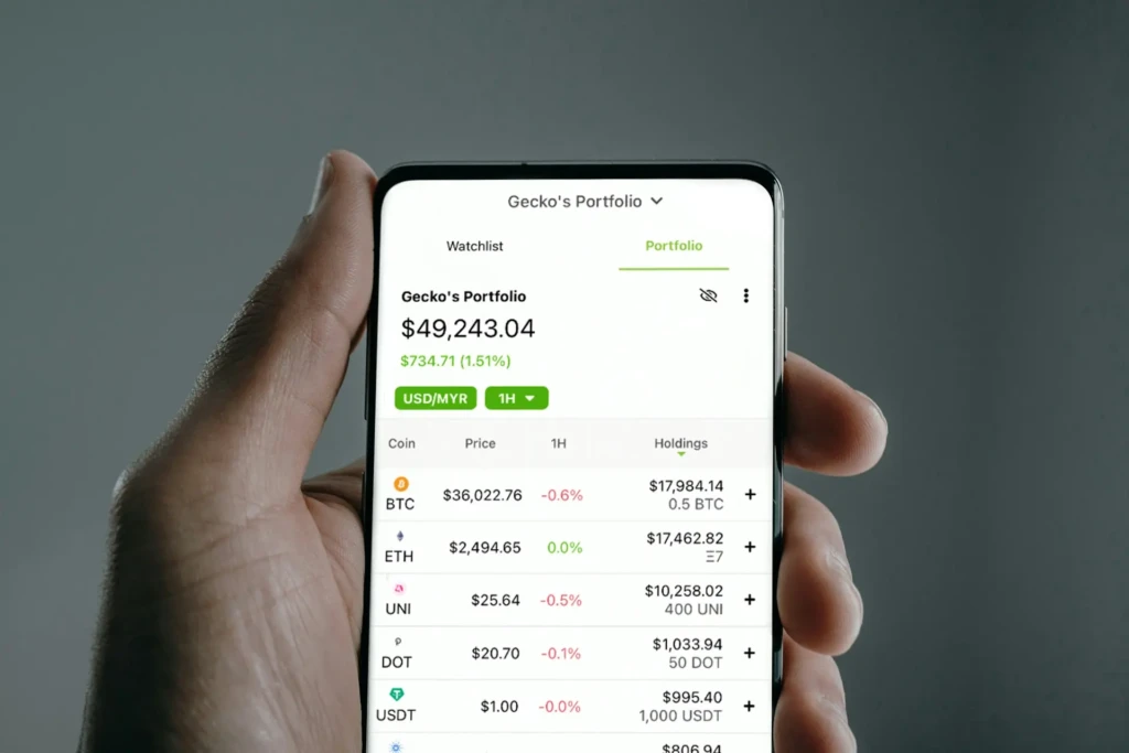 A hand holding a smartphone displaying “Gecko’s Portfolio” in Cash App, showing a total value of $49,243.04 (+$734.71), with holdings in BTC, ETH, UNI, DOT, and USDT, demonstrating its cryptocurrency portfolio tracking and real-time market data features.