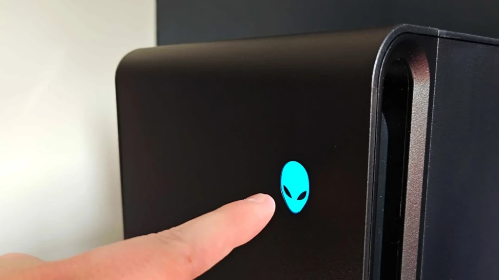 A close-up of a finger pressing the illuminated blue Alienware alien head logo on the front panel of a matte black desktop tower, showcasing the interactive power button and iconic branding in a minimalist studio setting.