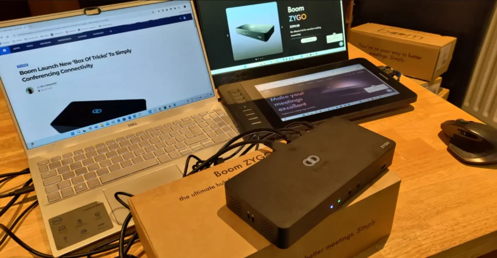 A workspace featuring a Dell laptop, tablet, and two external monitors connected to a black Boom ZYGO docking station resting on its retail box, with screens displaying product pages and reviews, demonstrating real-world multi-device connectivity for hybrid meetings and productivity.