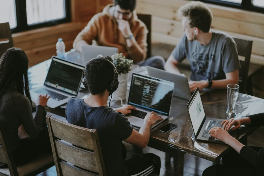 A candid overhead shot of four people collaborating around a wooden table with laptops, code editors open, headphones worn, and a small plant nearby, illustrating a real-world software development or startup team working in a modern, collaborative workspace.