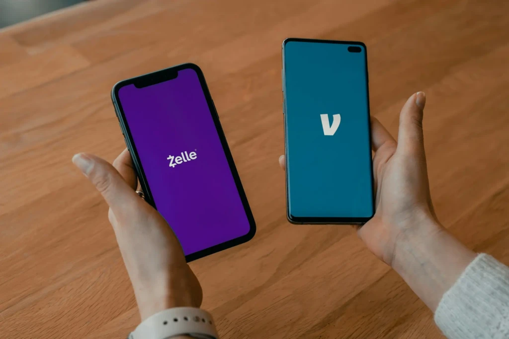 Two hands holding smartphones side by side on a wooden table: the left shows the Zelle app launch screen (purple background, white logo), and the right displays a competing app with a blue background and white “V” logo, suggesting a comparative review or feature contrast between payment services.