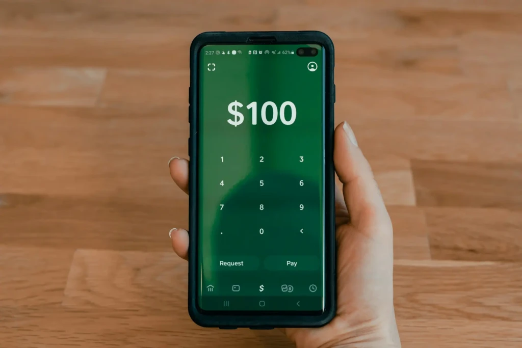 A hand holds a smartphone displaying a payment app with a green background showing "$100" and a numeric keypad.