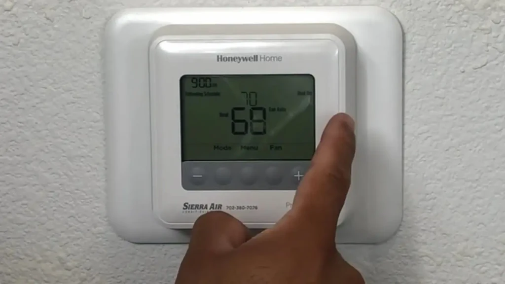 A hand adjusting the temperature on a Honeywell Home digital thermostat mounted on a textured wall, with the display showing “68”°F, “70” as setpoint, and “9:00 am,” illustrating manual control of heating/cooling in a residential setting.