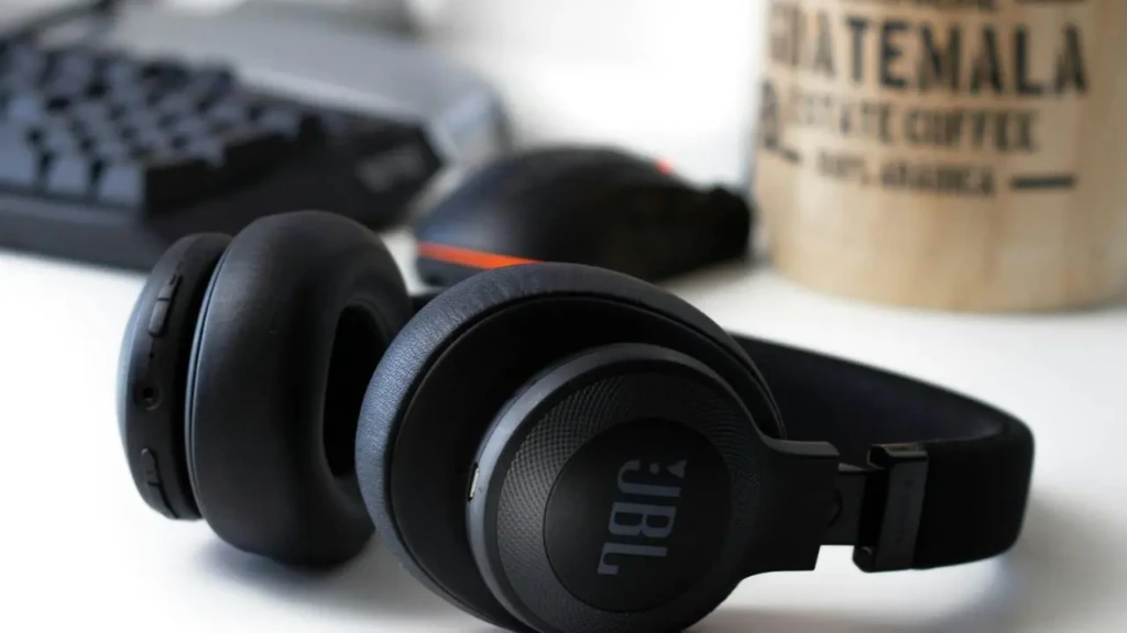 A pair of black JBL over-ear headphones resting on a white desk beside a keyboard, mouse, and a wooden coffee cup labeled “GUATEMALA COFFEE,” illustrating a modern workspace setup with integrated audio for productivity or leisure.