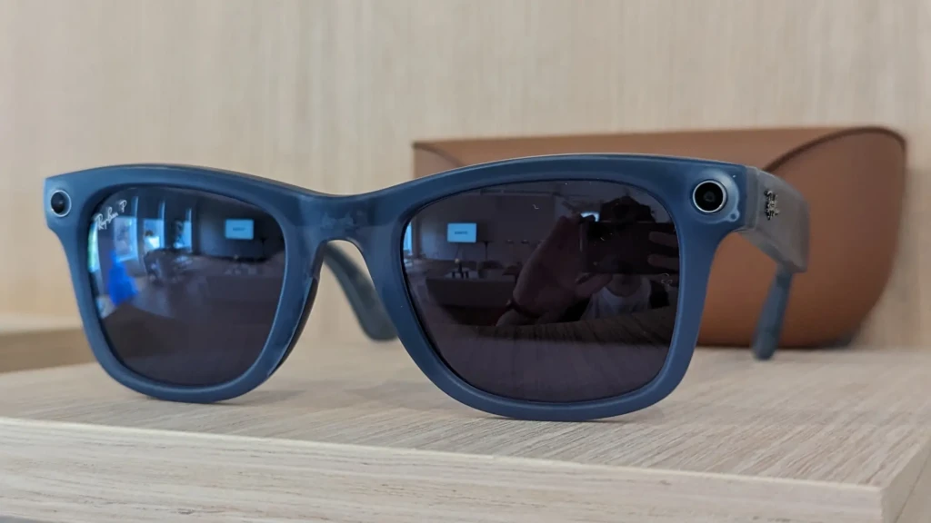 A pair of blue Ray-Ban Meta smart glasses placed on a light wood shelf, with visible cameras on the temples and reflections of a person taking the photo in the dark lenses, showcasing their design, build, and real-world placement alongside a brown case in the background.