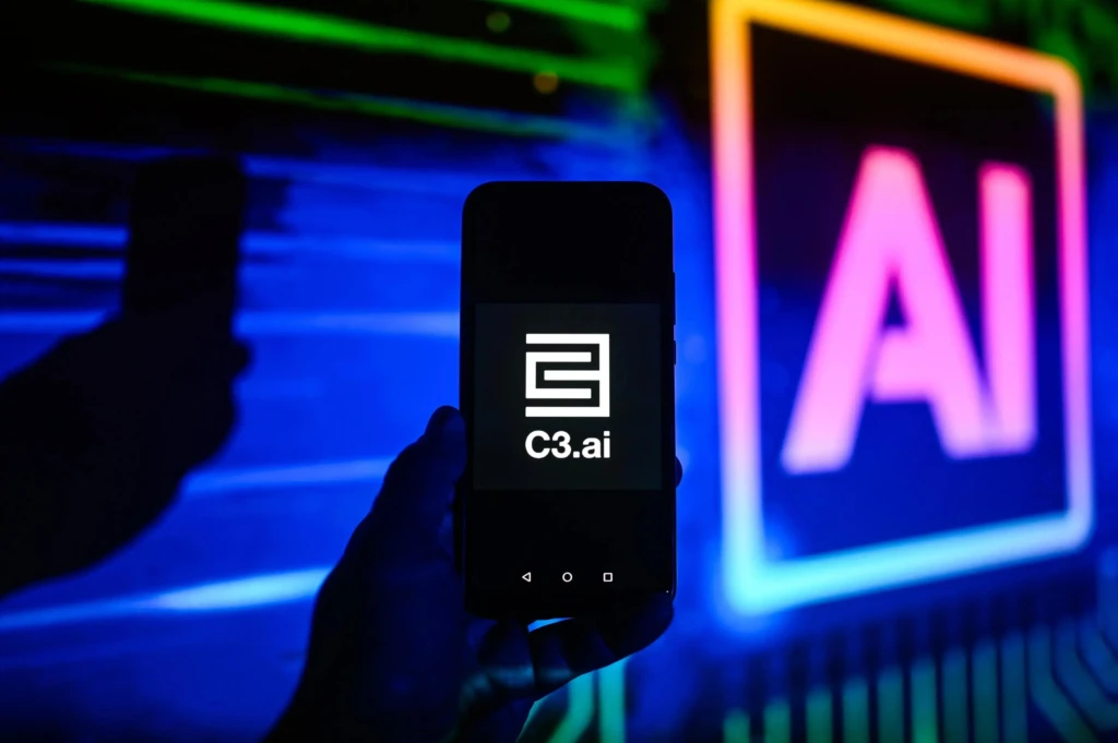 A hand holding a smartphone displaying the C3.ai logo, illuminated by colorful neon lights in the background.