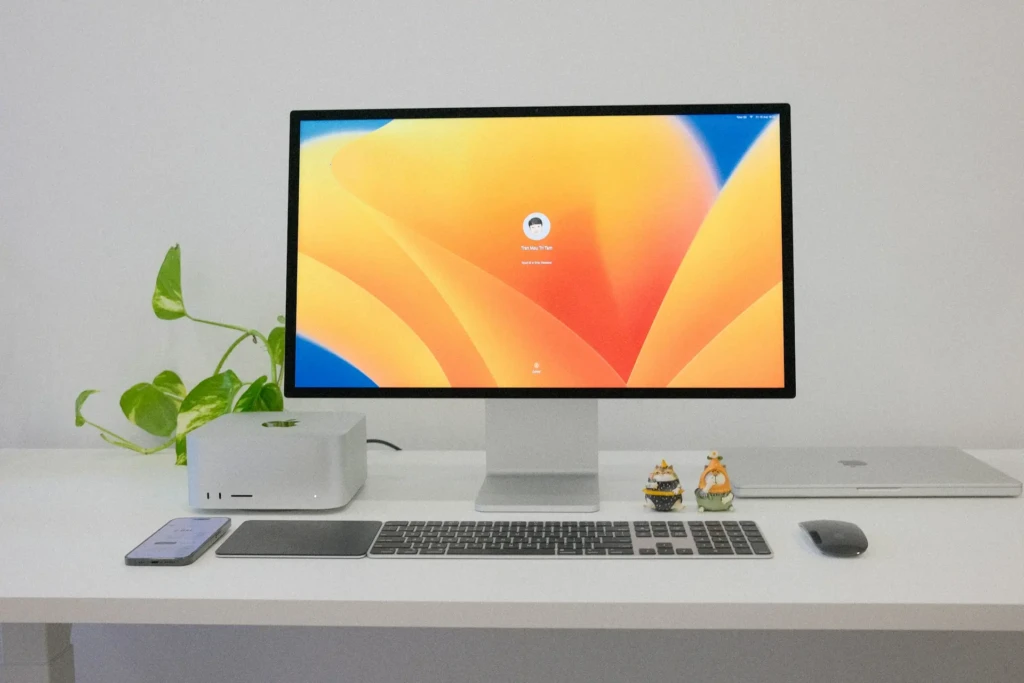 A minimalist Apple workspace featuring a Mac Studio with Apple logo, a large display showing the macOS login screen for user “Tran Mau Tri Tam,” a Magic Keyboard and Trackpad, an iPhone, a closed MacBook, two decorative figurines, and a green pothos plant, all arranged on a clean white desk against a neutral wall.