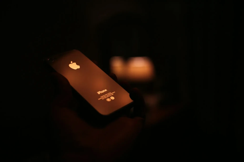 A hand holding a black iPhone in near-darkness, with the Apple logo and “iPhone” text faintly illuminated by warm ambient light from a blurred candle in the background, evoking a moody, intimate aesthetic that emphasizes the device’s silhouette and reflective surface.