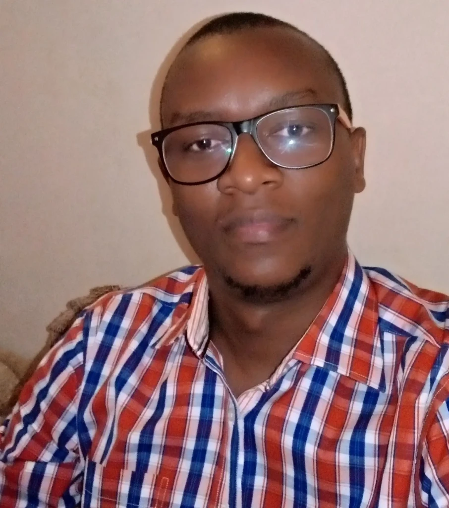 A close-up portrait of a man with short hair, glasses, and a light beard, wearing a red-, blue-, and white-striped button-down shirt, seated against a plain beige wall, capturing a direct, neutral expression in natural indoor lighting.
