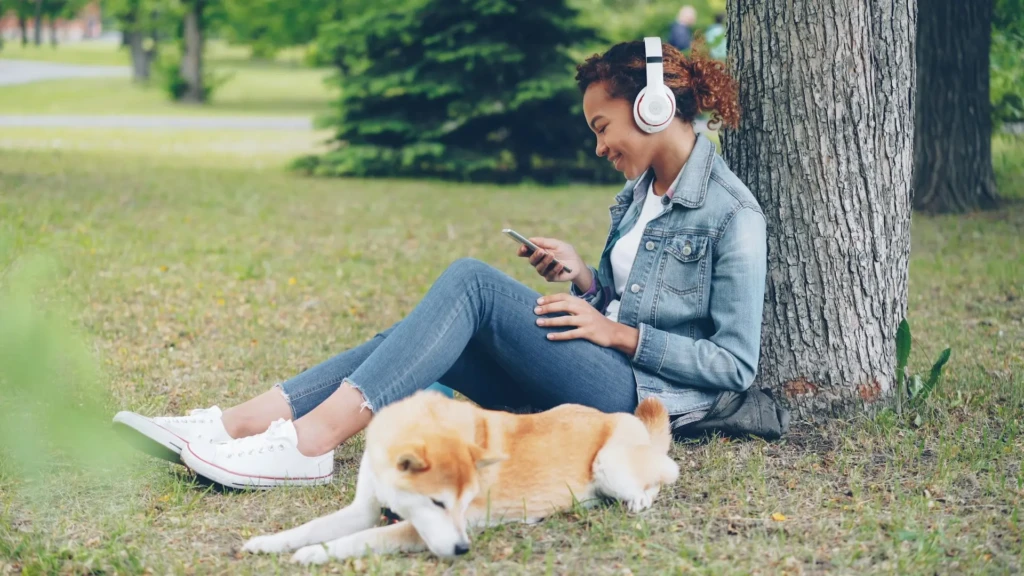 A young woman wearing headphones sits against a tree using her phone, while a dog lies on the grass beside her in a park.