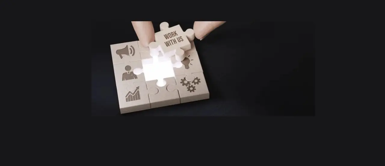 A hand places a puzzle piece labeled "WORK WITH US" into a puzzle featuring various business icons on a dark background.