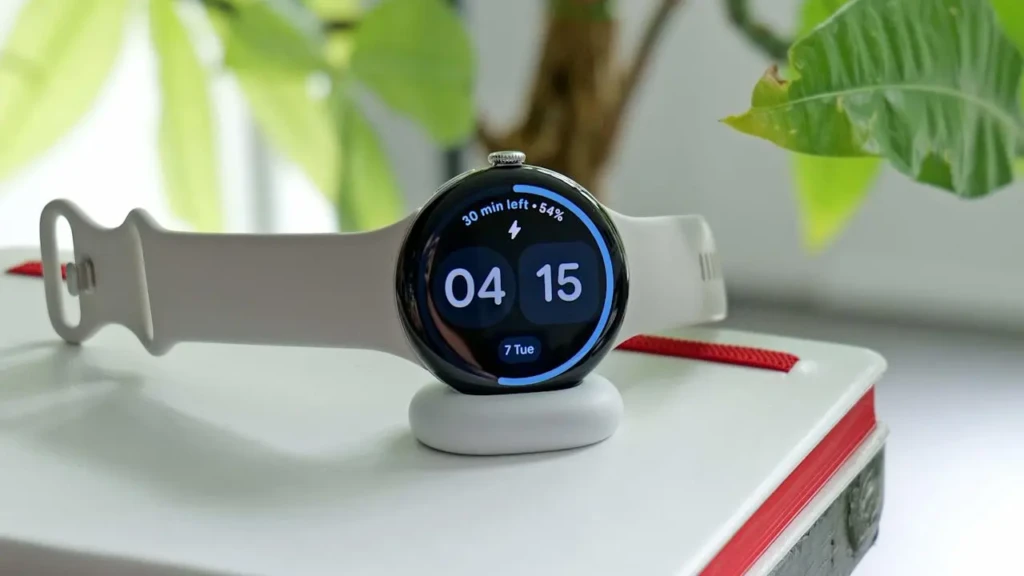 A white Google Pixel Watch resting on a charging dock atop a book, with its screen showing “04:15”, “7 Tue”, and a circular progress bar indicating “30 min left • 54%,” set against a soft-focus plant background, highlighting sleek design and seamless charging.