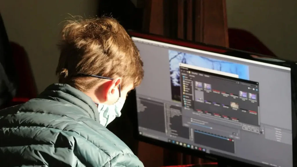 Person wearing a green jacket and mask working on a computer with photo editing software open on the monitor.