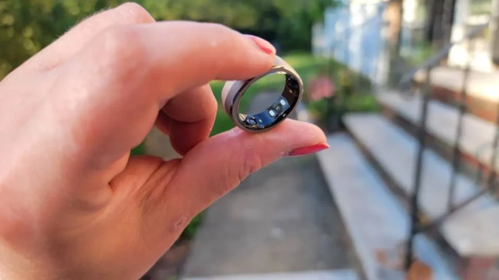 A close-up of a hand with pink nail polish holding a metallic smart ring between thumb and index finger, showcasing its slim profile and internal sensors, with a blurred outdoor sidewalk and greenery in the background, illustrating real-world portability and everyday wearability.