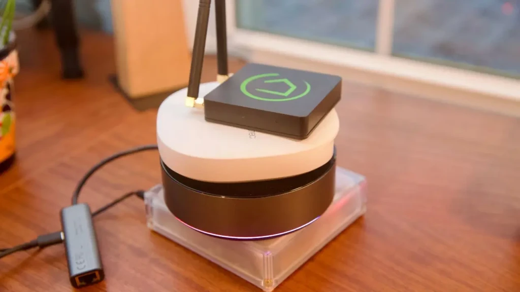 A black smart streaming device with a green logo stacked atop a white circular hub and a black base unit, all placed on a clear acrylic stand with an Ethernet adapter connected, highlighting layered connectivity and modular smart home setup on a wooden surface.