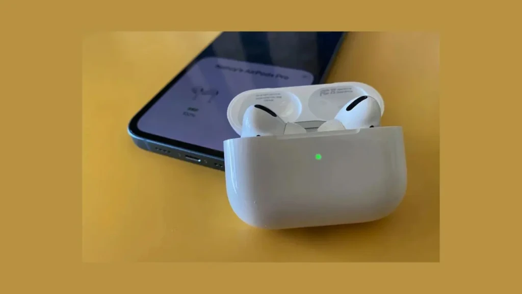 White wireless earbuds in an open charging case with a glowing green indicator, placed next to a smartphone showing a battery level screen on a yellow surface.
