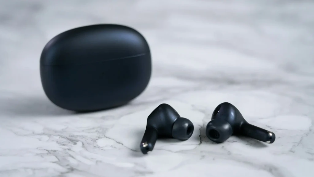 Two matte black wireless earbuds placed on a white marble surface beside their closed oval-shaped charging case, highlighting minimalist aesthetics, ergonomic shape, and premium material finish in a clean product presentation.
