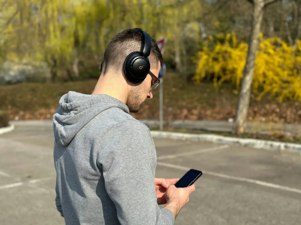 A person wearing headphones and a gray hoodie looks at a smartphone outdoors, surrounded by trees and bright yellow shrubbery, conveying a relaxed mood.