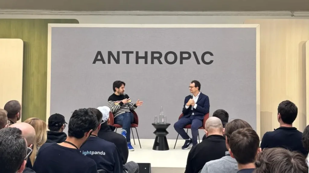 Two speakers seated on a stage in front of a large gray backdrop with the “ANTHROPIC” logo, addressing an audience in a live event setting, capturing a public discussion or presentation by Anthropic representatives.