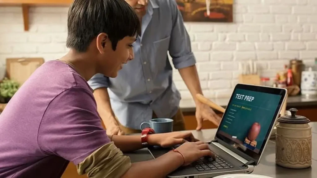 A young student using a laptop displaying “TEST PREP” while an adult mentor leans in to assist, in a home kitchen setting with mugs and decor, illustrating guided, technology-supported learning and homework help.