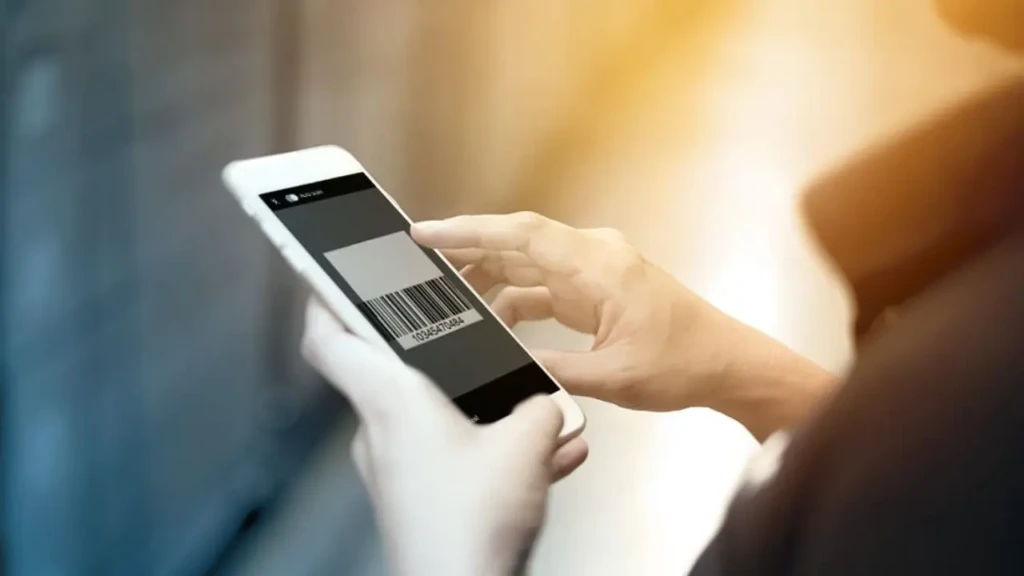 A close-up of hands holding a white smartphone displaying a barcode-scanning interface, with a finger poised to tap the screen, illustrating real-time product scanning for nutritional data entry in a health or fitness app.