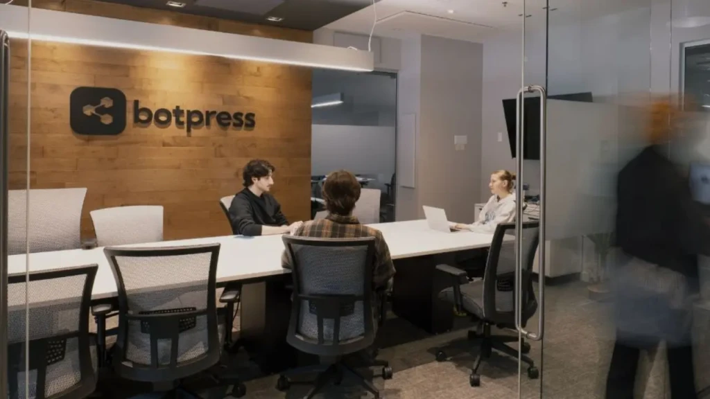 A modern conference room with a wooden accent wall features the "botpress" logo. Three people are seated around a table, engaging in discussion.