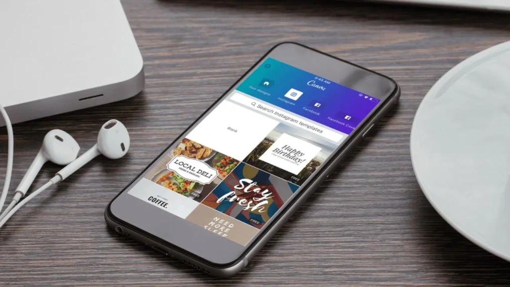 A smartphone on a wooden desk showing the Canva app interface with Instagram template previews (e.g., “Happy Birthday!”, “Stay fresh”), next to earbuds, a laptop corner, and a ceramic plate, illustrating social media content creation using mobile design tools.